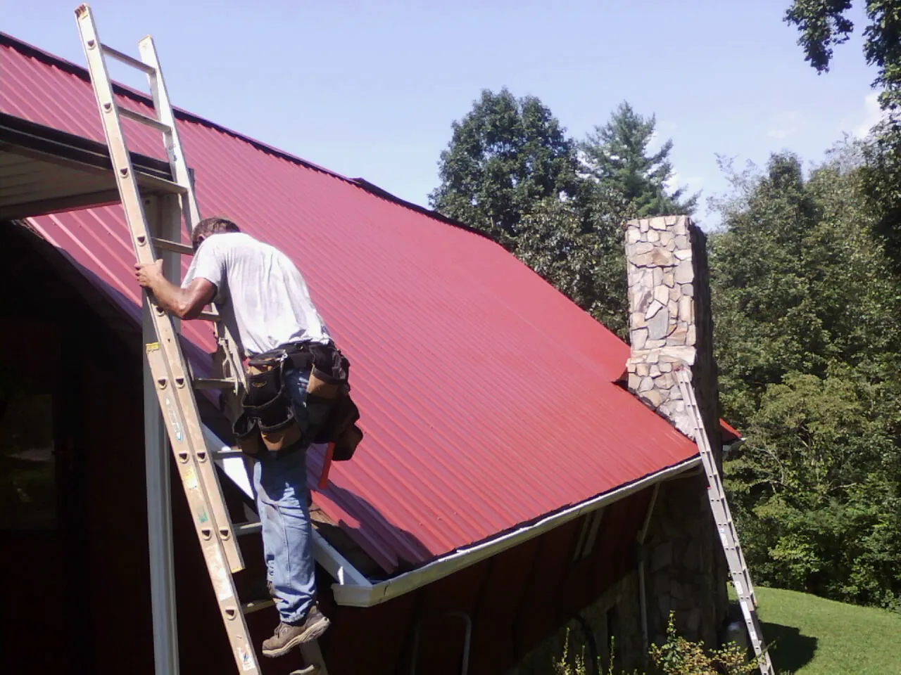 Roofer on a ladder beside a red metal roof and stone chimney — Soffit & Fascia Repair by Roofing Brothers in Custer City